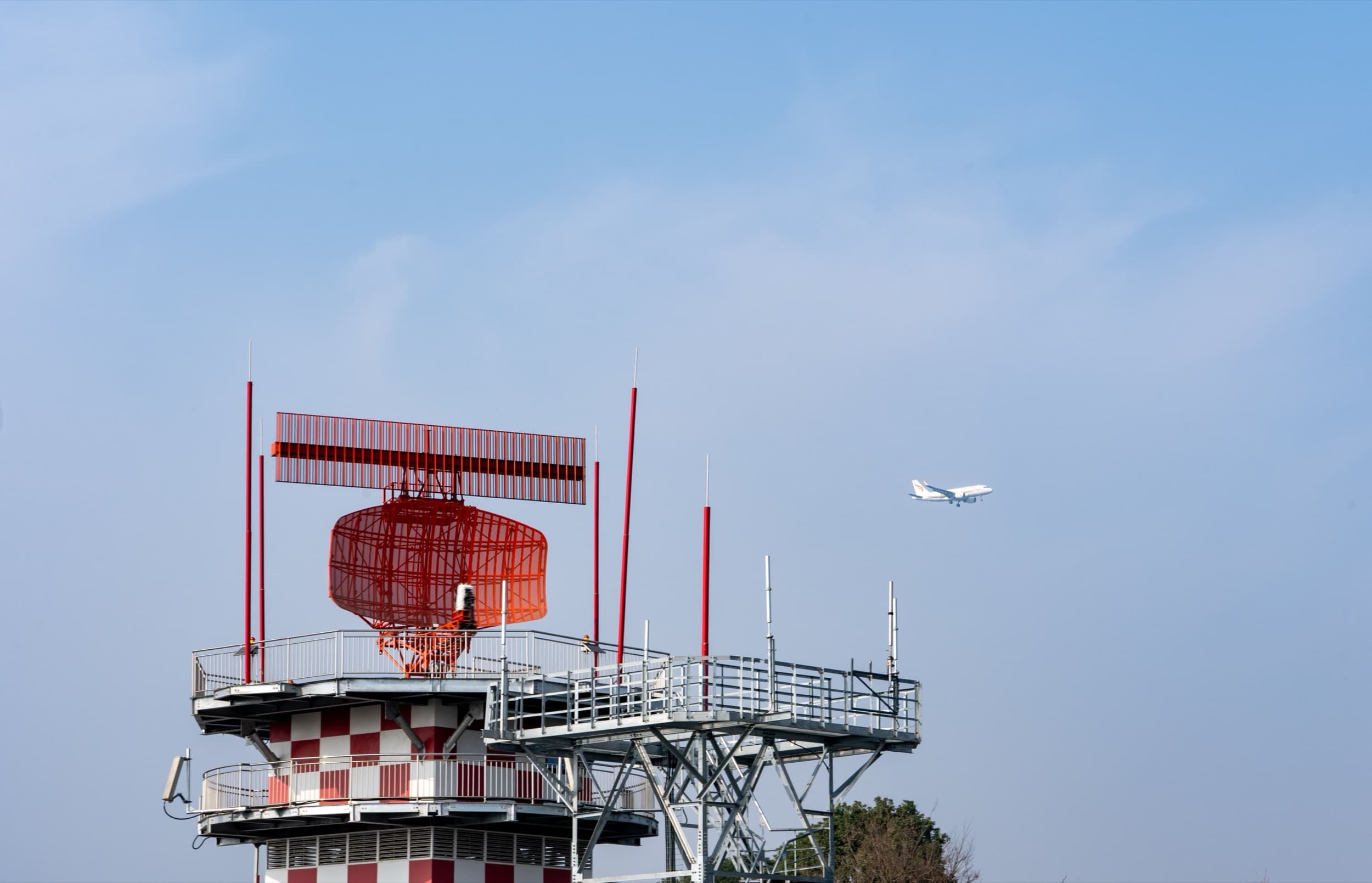 Rot-weißer Radarturm eines Flughafens, im Hintergrund ein Flugzeug im Anflug