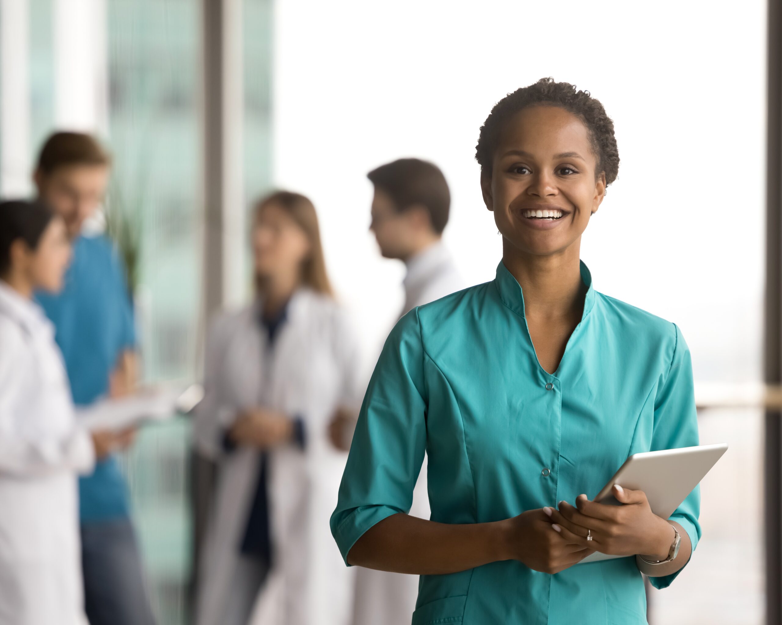 Happy young African American doctor woman holding tablet Ein leerer Raum mit vielen großen Fenstern wird sehr unscharf dargestellt. Es handelt sich wohl um ein Krankenhaus, was zu E-Health passt