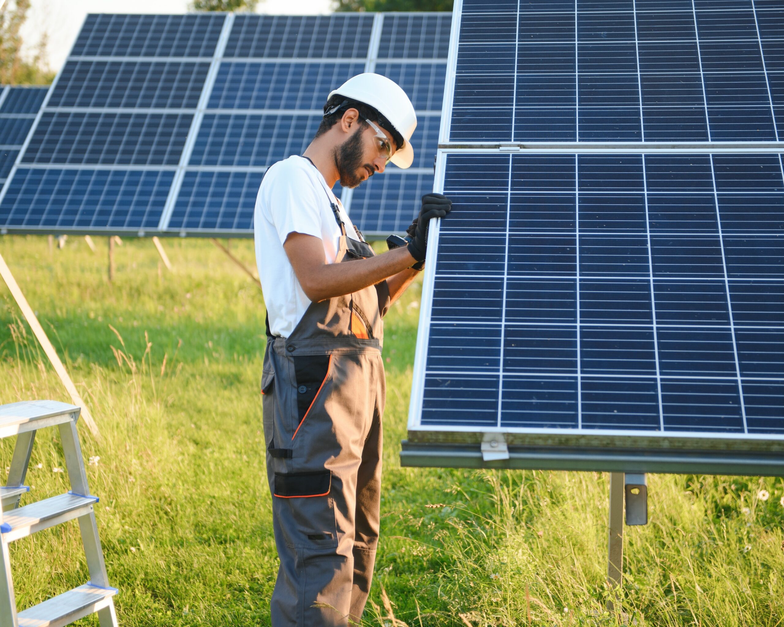 Male arabian engineer in helmet and brown overalls checking resistance in solar panels outdoors. Indian man working on station. Eine Photovoltaik-Anlage auf einem Dach. Aus dem Hintergrund scheint die untergehende Sonne schräg auf das Dach. Die Solarpaneele stehen symbolisch für die Versorgungswirtschaft