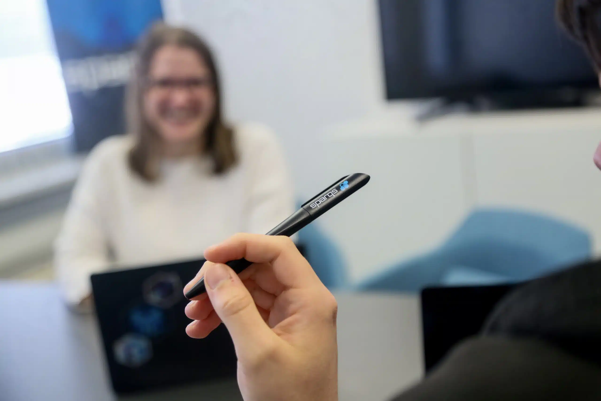 Im Vordergrund hält eine Person einen schwarzen Kugelschreiber, auf dem "sparqs" steht. Im unscharfen Hintergrund sitzt eine lachende Frau. Das ist über uns.